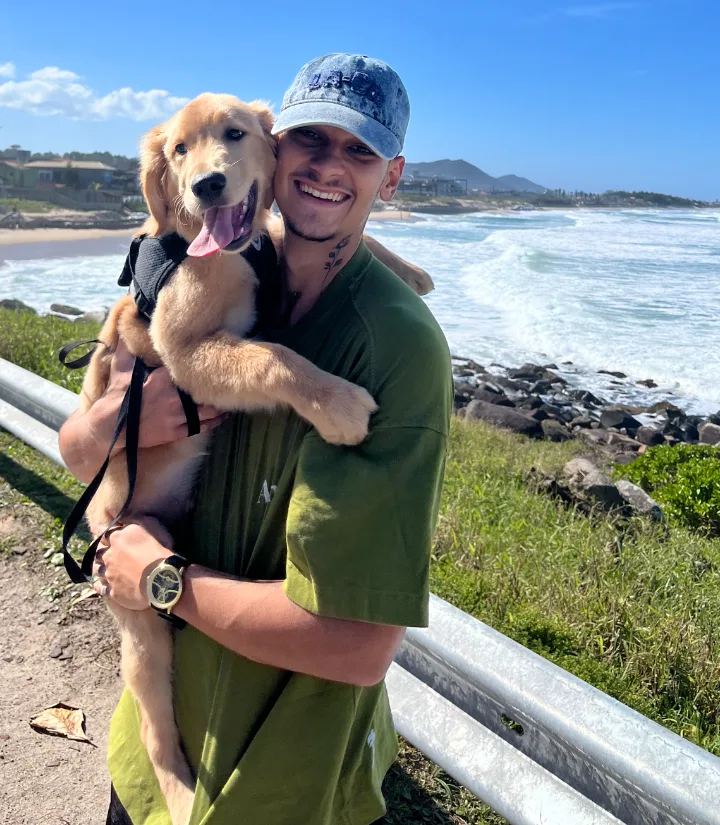 Heric sorrindo na praia, abraçado com um cachorro golden retriever